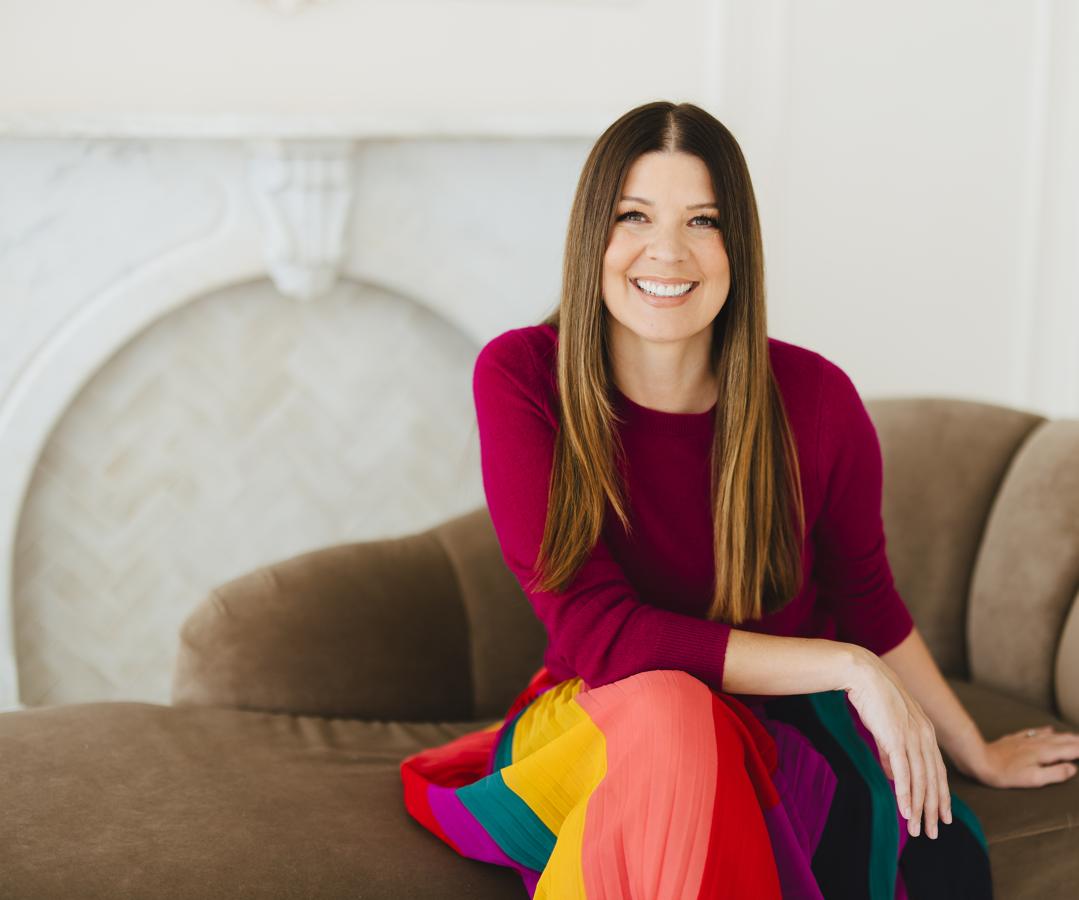 Dr. Stephanie Helfand seated on a sofa, smiling, in a magenta sweater and a colorful striped skirt.