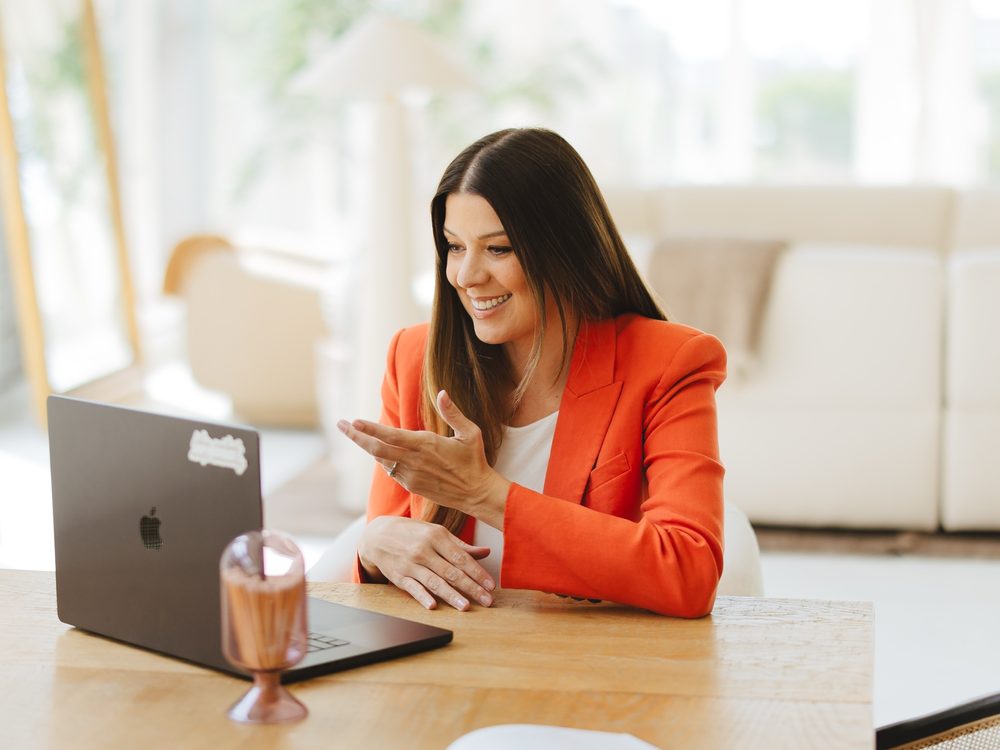 Dr. Stephanie Helfand in an orange blazer, smiling and talking with someone on a video call at a sunny table.